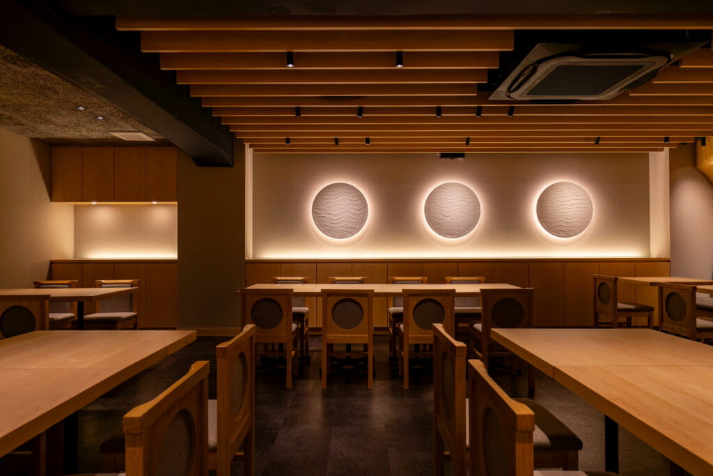 和モダンなデザインのレストラン内観。木製の天井と家具、壁には間接照明で照らされた円形のアートが3つ並んでいる。